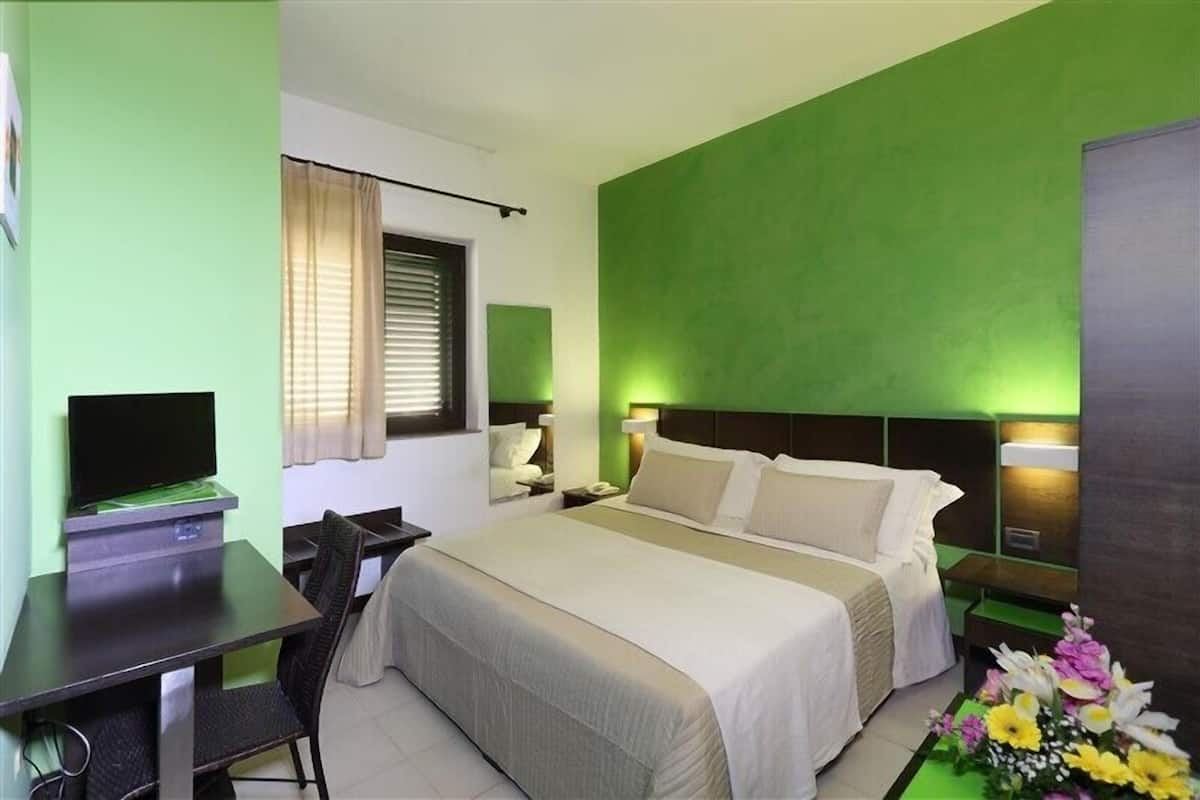 Une chambre d’hôtel avec un lit, un bureau avec un ordinateur, une chaise, une télévision et une fenêtre avec des rideaux.
