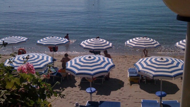 Een strand met gestreepte parasols en ligbedden, een zeilboot in de verte, en een helder uitzicht op de zee.