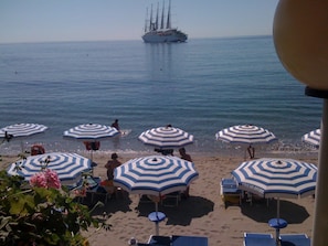 A beach with striped umbrellas and lounge chairs, a sailboat in the distance, and a clear view of the sea.