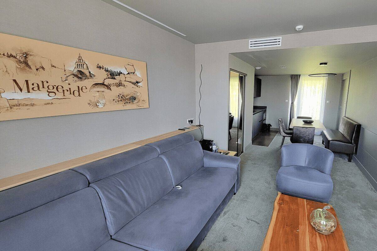Uma sala de estar moderna com um sofá cinzento, uma mesa de centro de madeira e uma área de jantar com mesa e cadeiras. Uma grande obra de arte está visível na parede.