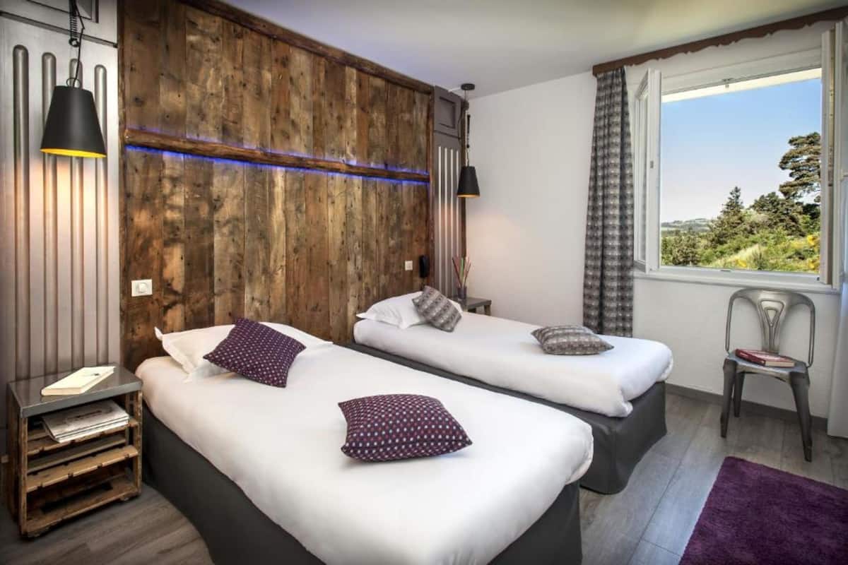 Quarto com duas camas, parede decorativa em madeira, janela com vista e uma pequena mesa com cadeira.