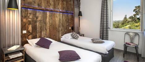 A room with two beds, a wooden accent wall, a window with a view, and a small table with a chair.