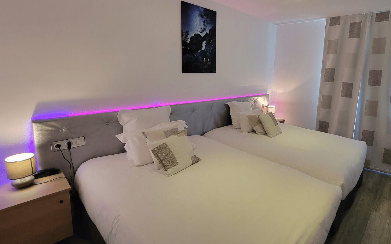 A double bed with white bedding and a headboard with a purple light strip. There is a nightstand with a lamp and a power outlet. A painting hangs on the wall above the bed.