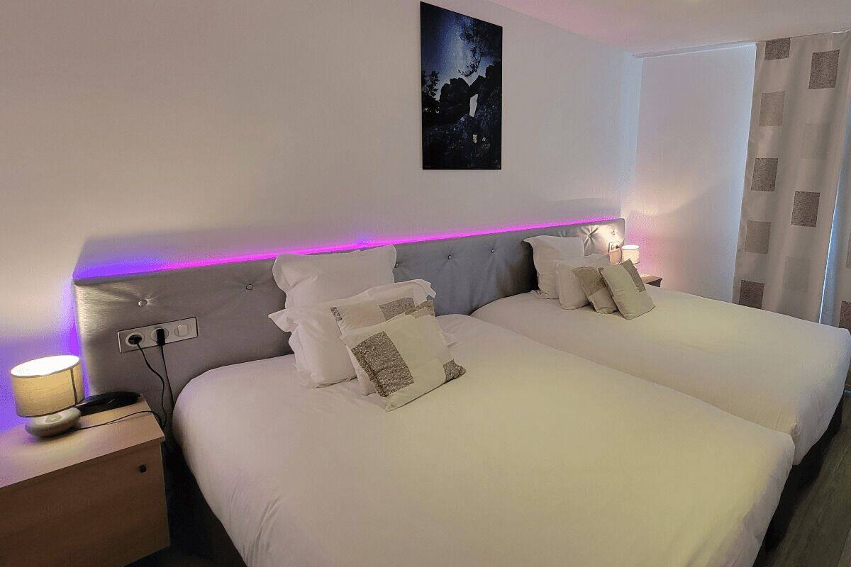 Cama de casal com roupa de cama branca e cabeceira com fita de luz roxa. Há uma mesa de cabeceira com abajur e tomada. Um quadro está pendurado na parede acima da cama.