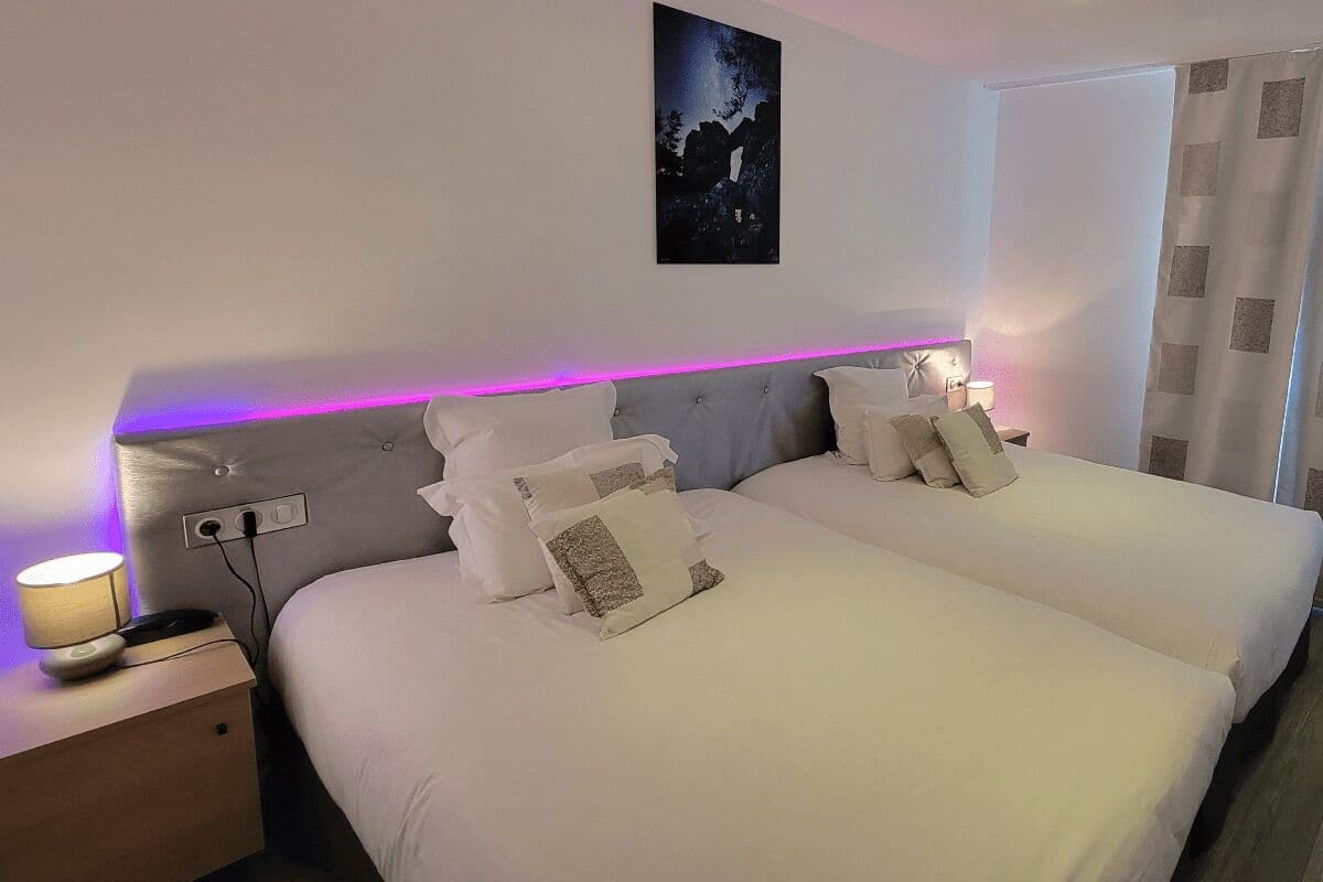 Cama doble con ropa de cama blanca y cabecera con una tira de luz púrpura. Hay una mesita de noche con lámpara y toma de corriente. Un cuadro cuelga en la pared encima de la cama.