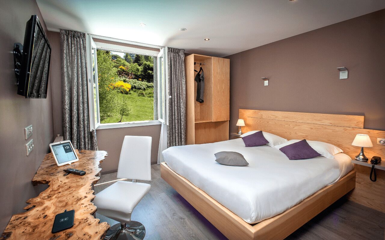 A modern hotel room with a large bed, a wooden desk, a chair, a TV, and a view of greenery outside.