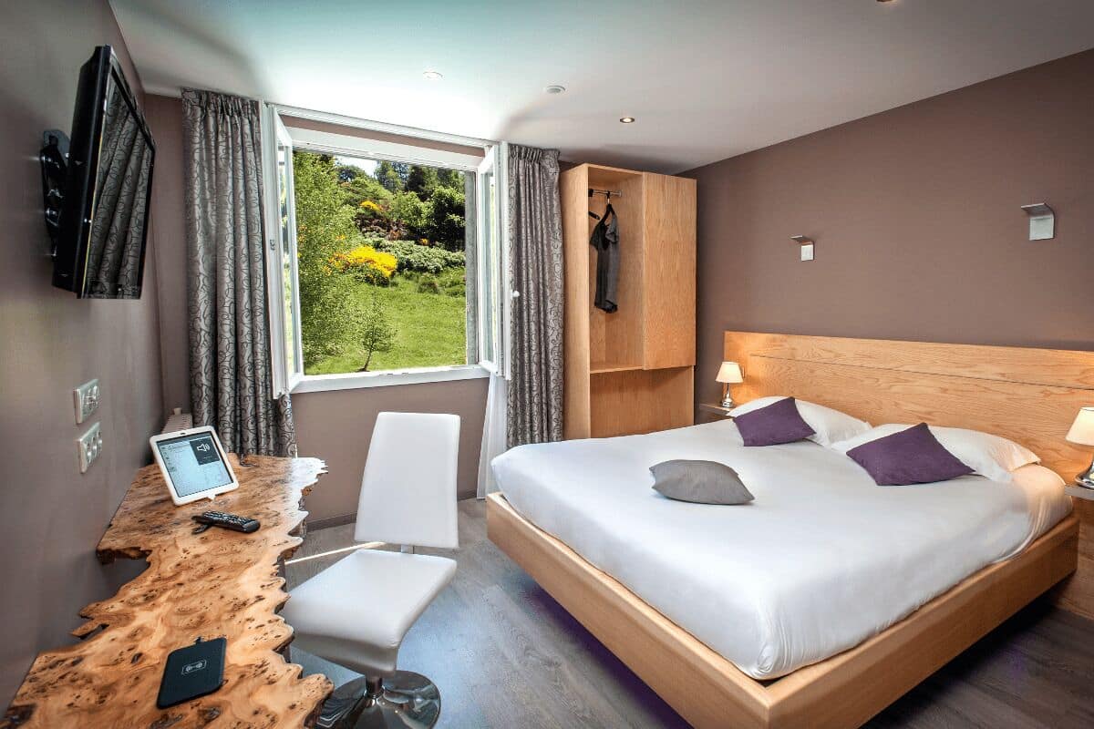 Quarto de hotel moderno com uma cama grande, uma escrivaninha de madeira, uma cadeira, uma TV e vista para o verde lá fora.