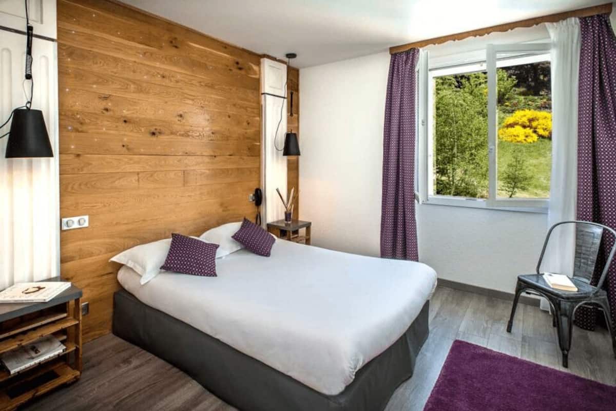 Quarto com parede de madeira, cama com roupa de cama branca, uma cadeira, janela com cortinas e uma pequena mesa com luminária.