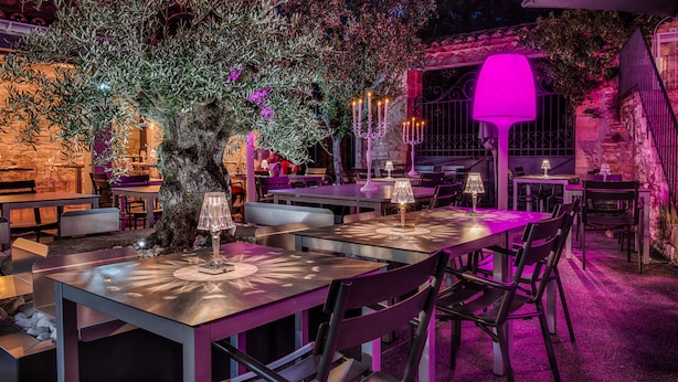An outdoor dining area with tables and chairs, a large tree, and a purple-lit lamp post. - Le Mas du Terme (Barjac)