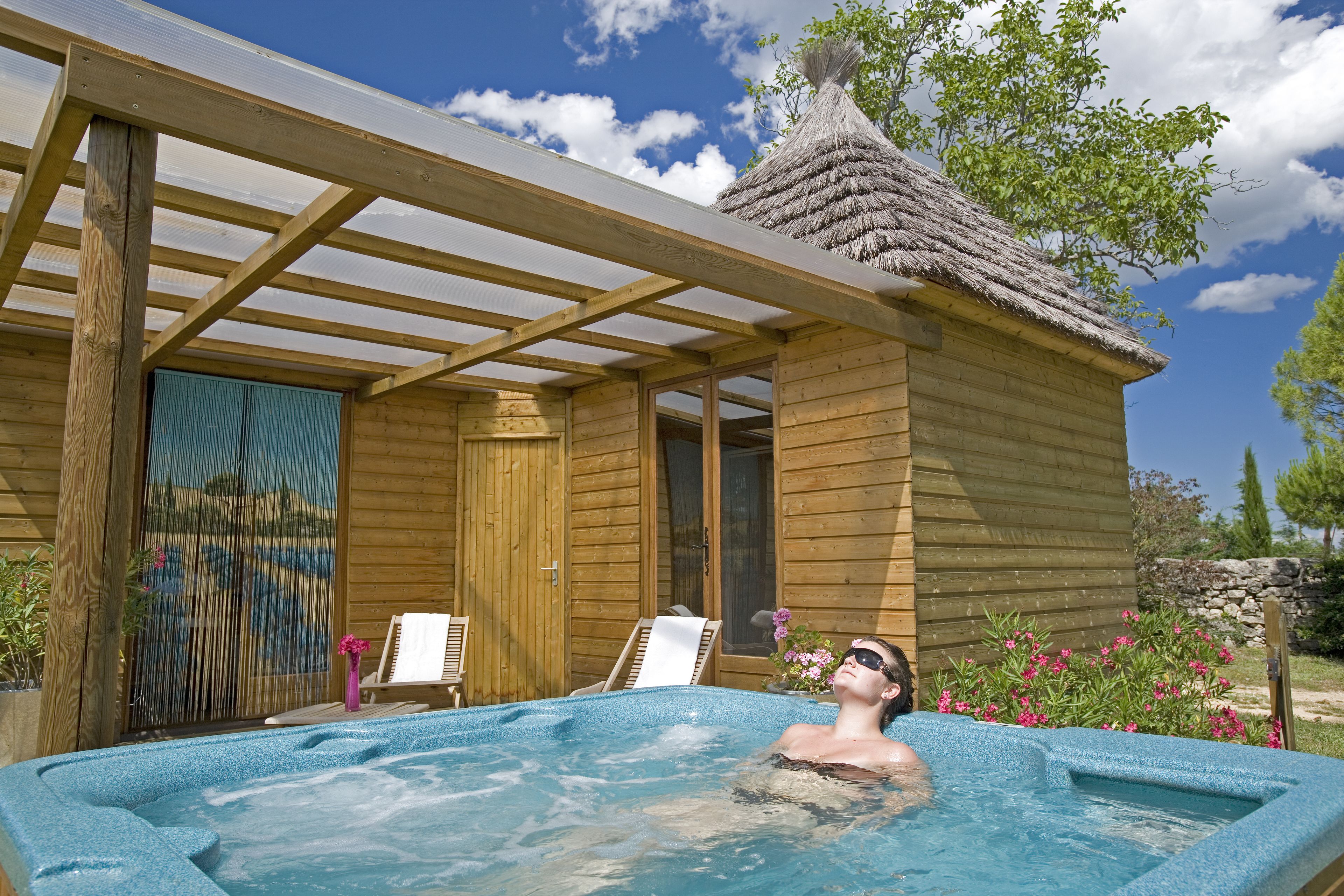 A wooden cabin with a hot tub, wooden deck, and a thatched-roof structure.