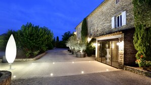 A stone building with a paved driveway, potted plants, and a well-lit entrance. - Le Mas du Terme (Barjac)
