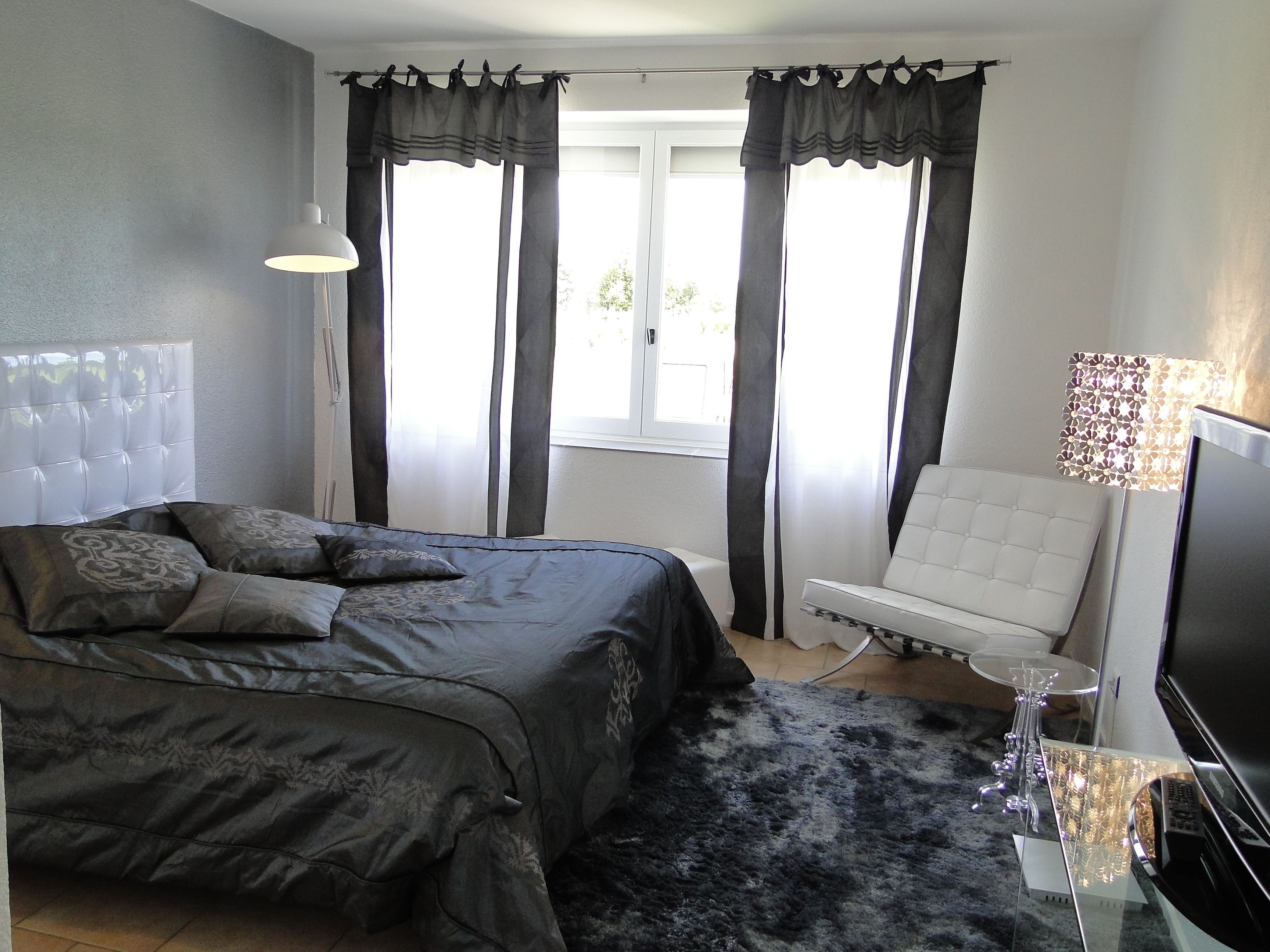 Un dormitorio con cama, una silla, televisor y ventana con cortinas.