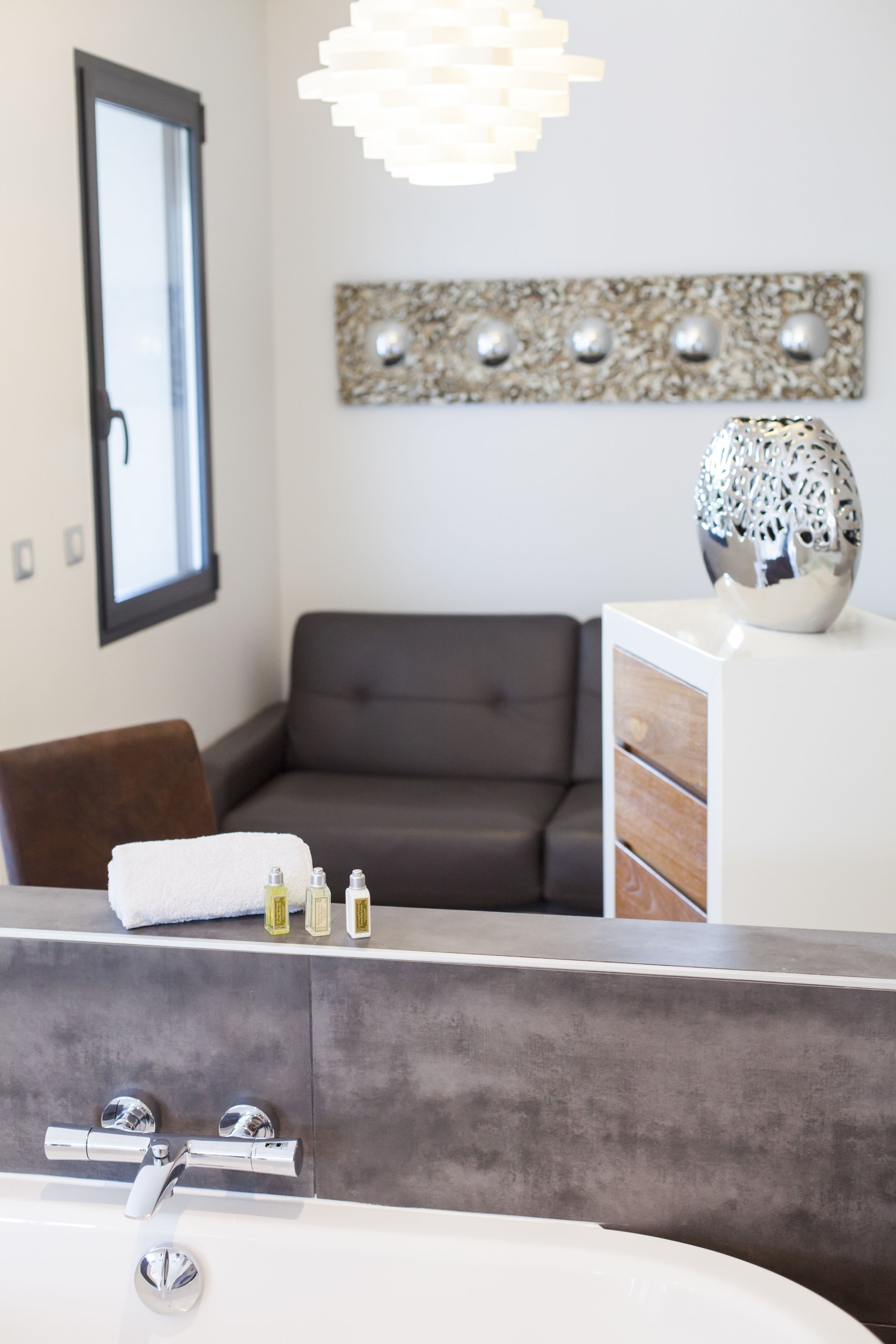 A modern bathroom with a bathtub, a gray sofa, a decorative wall piece, and a white cabinet with toiletries.