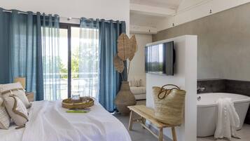 A modern bedroom with a large bed, a TV, a bathtub, and a view of greenery outside.