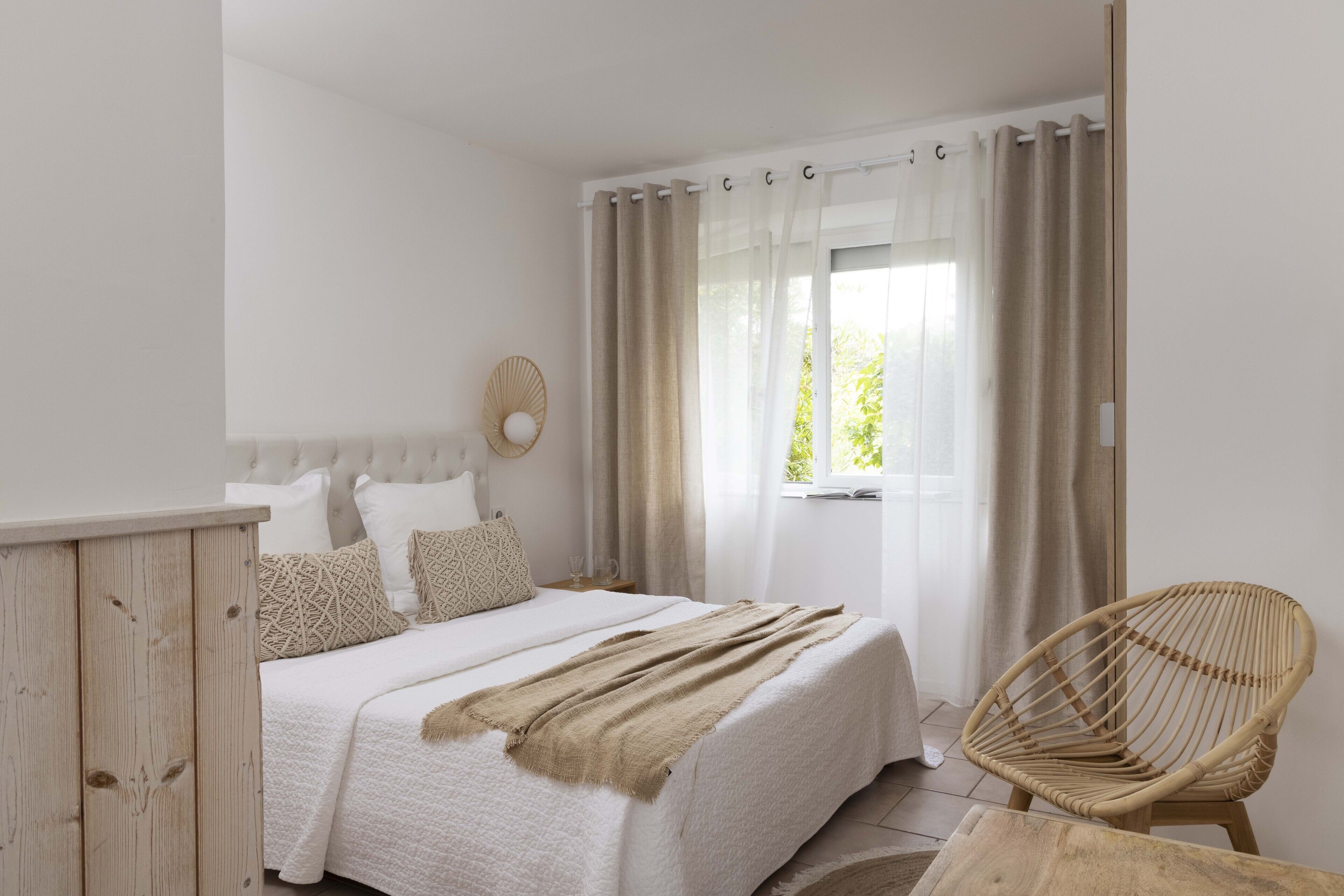 Quarto com cama, guarda-roupa de madeira, cadeira de vime e janela com cortinas.