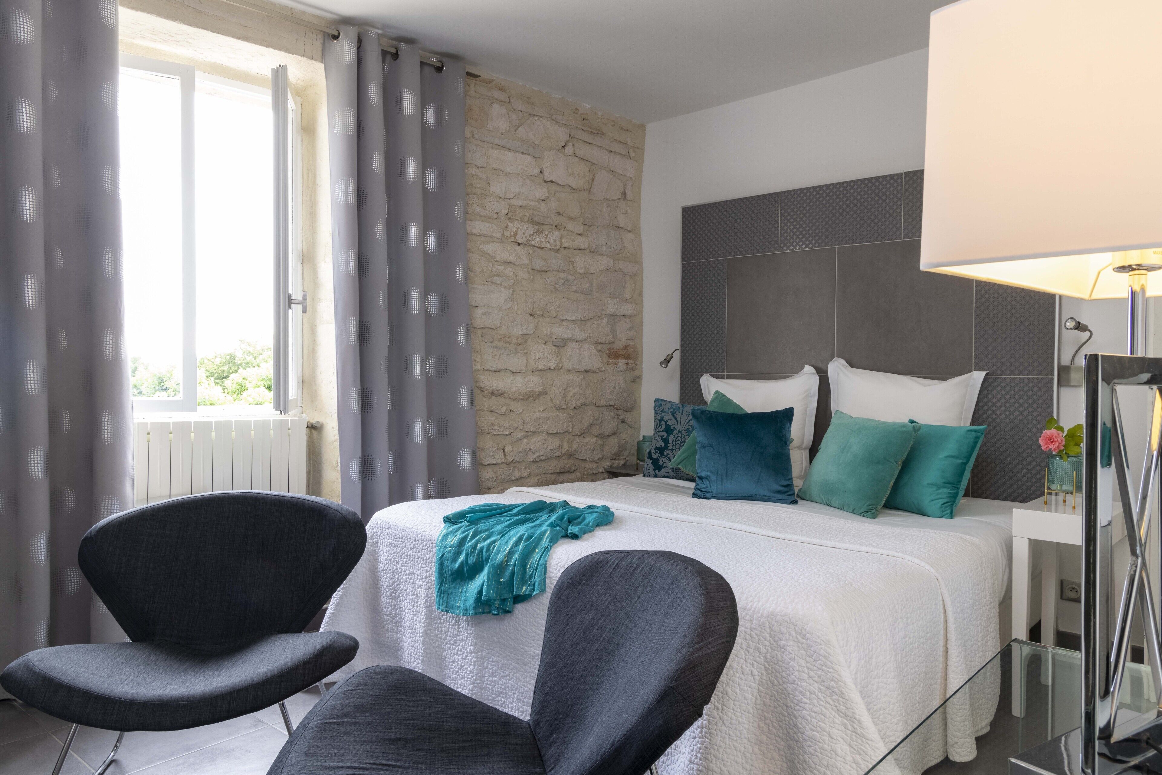 Un dormitorio moderno con una pared de acento de piedra, una cama grande con ropa de cama blanca y almohadas turquesa, una mesita de noche con lámpara, y vistas a zonas verdes al exterior.