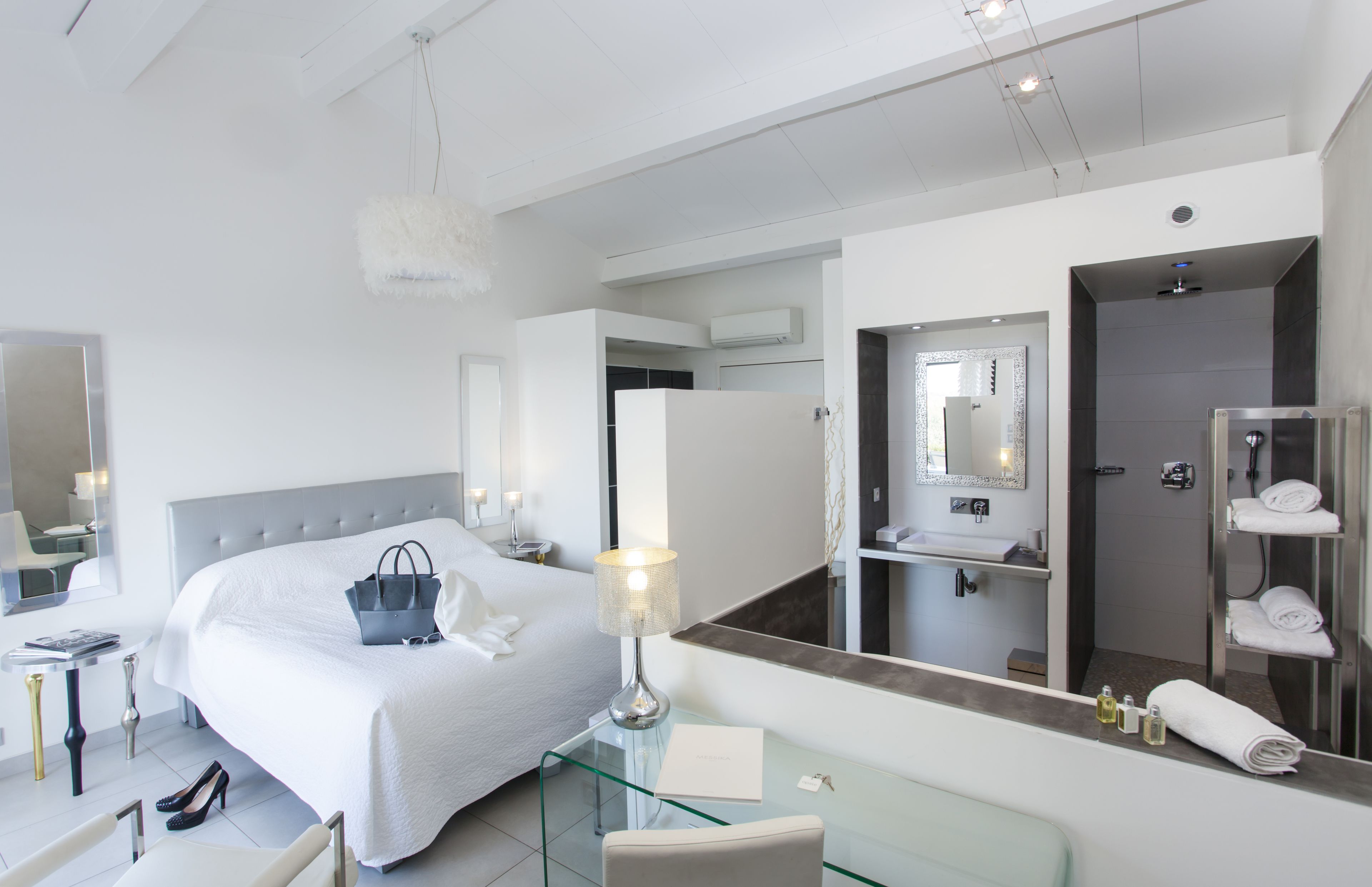 A modern hotel room with a large bed, a glass coffee table, a sitting area, a bathroom with a shower, and a large mirror.