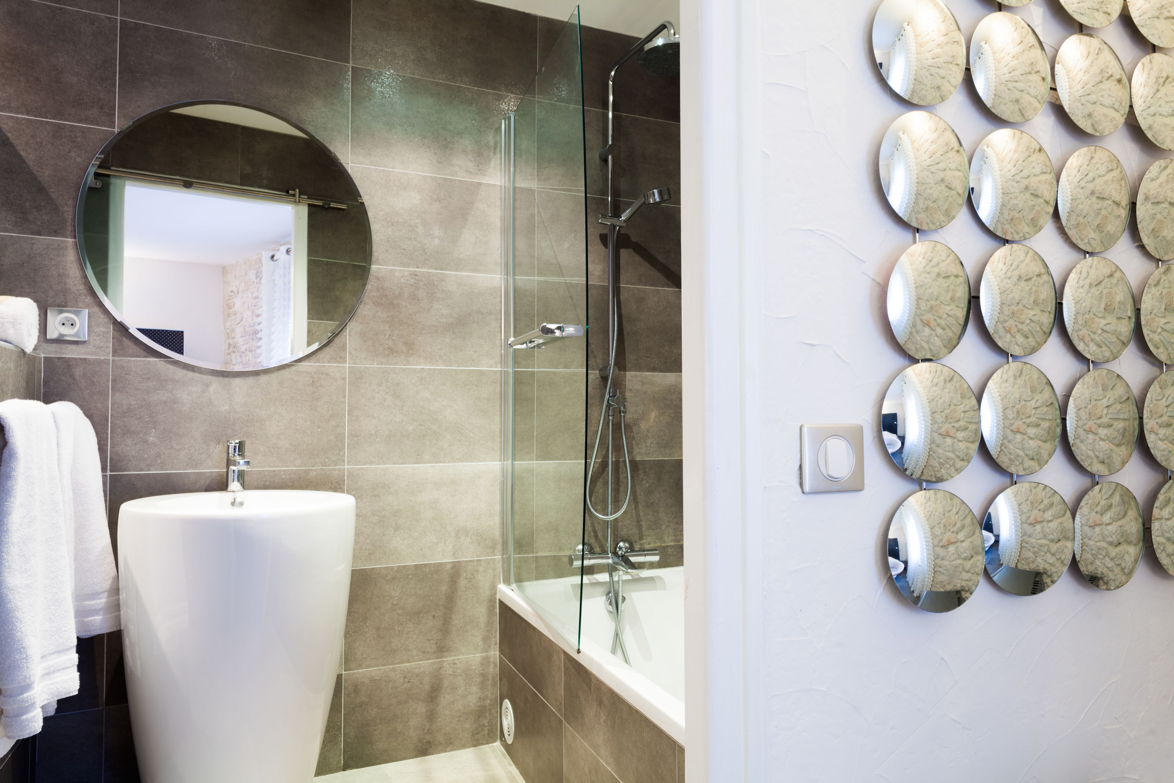 A bathroom with a round mirror, a sink, a shower, and a wall-mounted mirror arrangement.