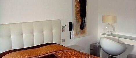Un dormitorio con un cabecero capitonado, una cama con colcha estampada, un cuadro en la pared, una lámpara de noche y una mesita con lámpara.