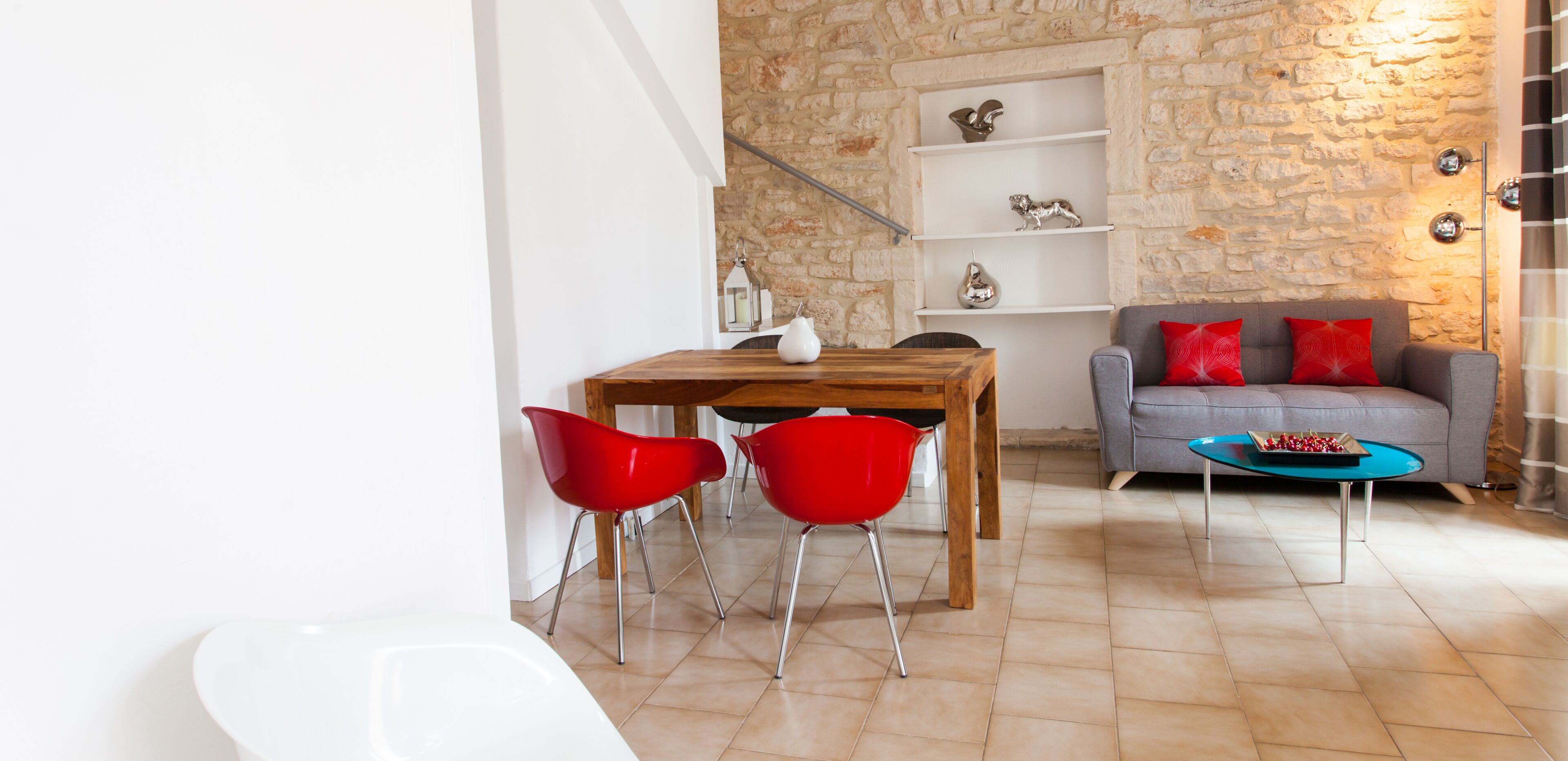 A dining area with a wooden table and red chairs, a grey sofa with red cushions, and a blue coffee table.