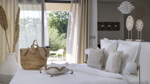 A bedroom with a large bed, white bedding, a bedside table with a lamp, and a view of greenery outside. - Le Mas du Terme (Barjac)
