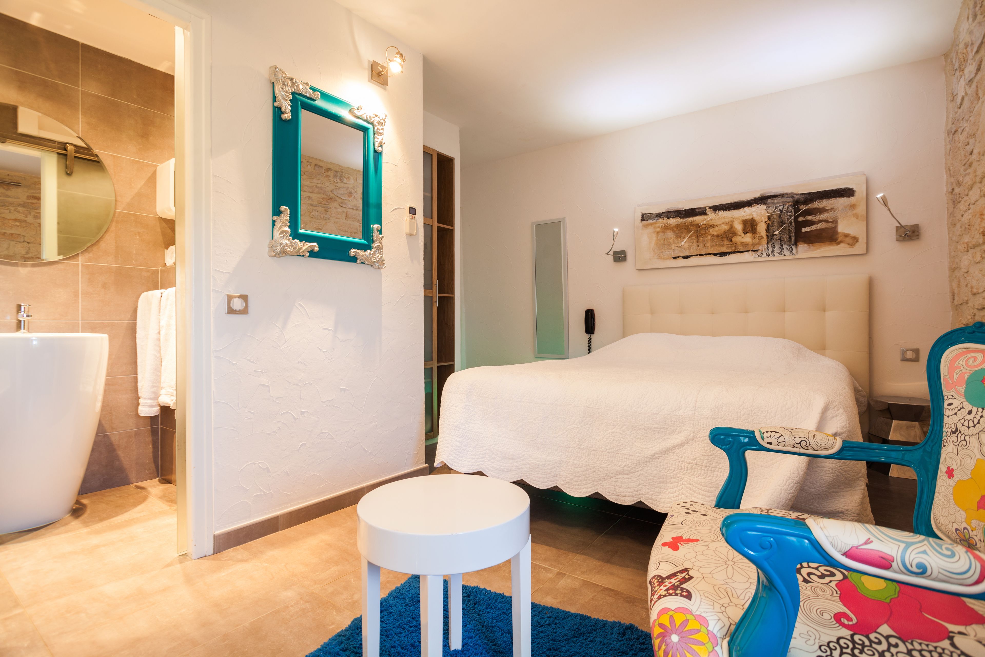 Un dormitorio con una cama, una silla, un taburete pequeño, un espejo y una obra de arte colgada en la pared.