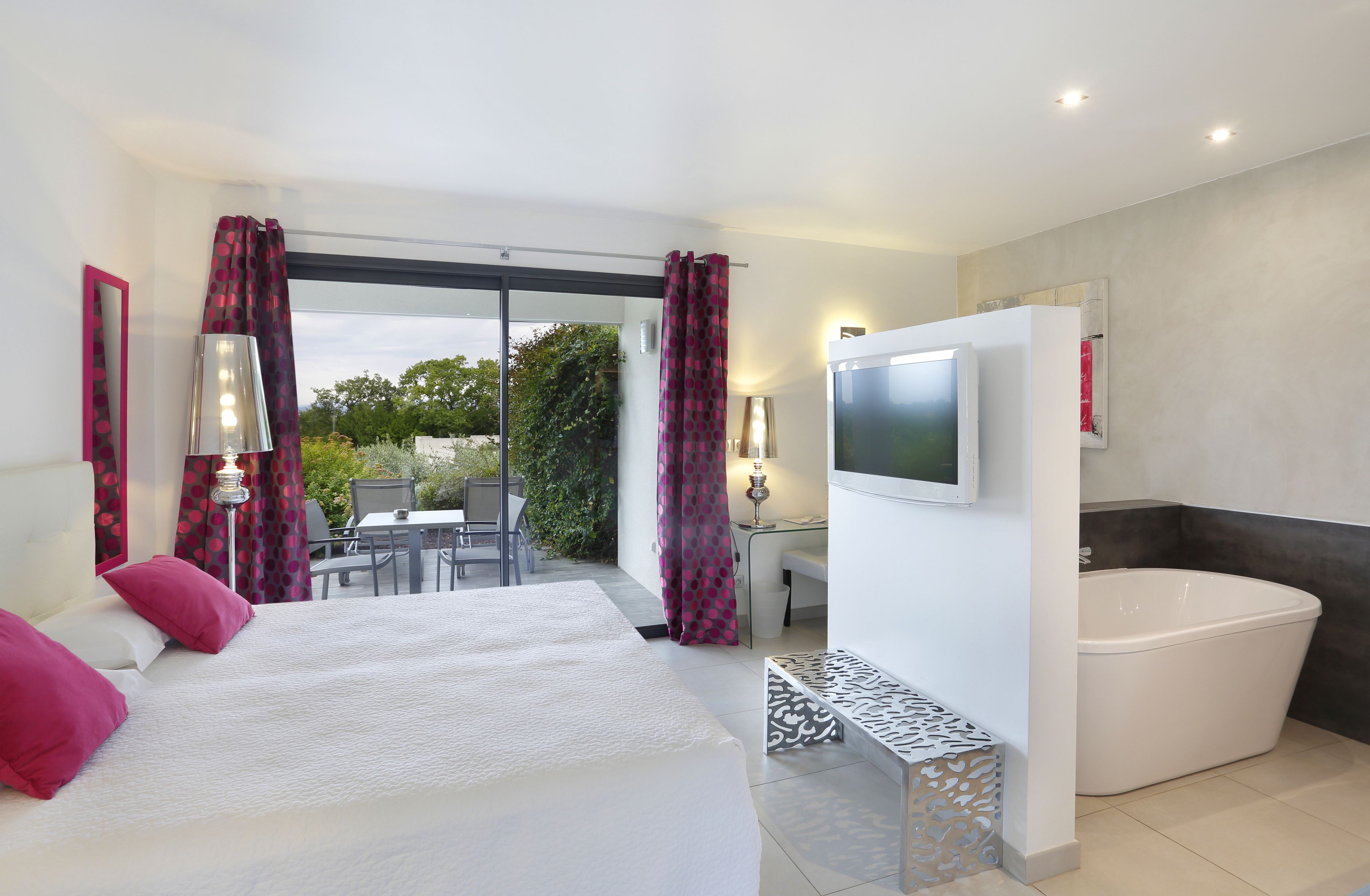 A modern bedroom with a large bed, a TV, a bathtub, and a balcony with a table and chairs.