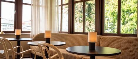 A seating area with round tables and chairs, large windows with curtains, and a view of greenery outside.
