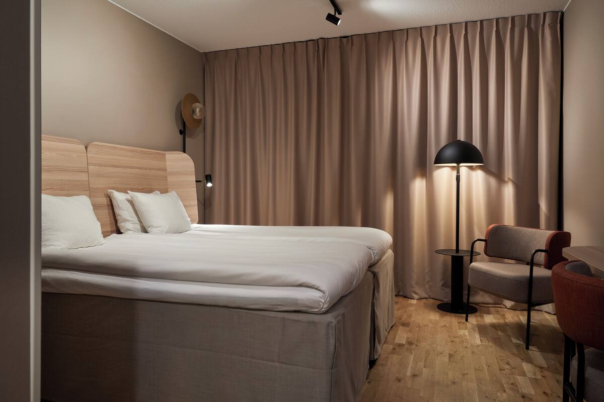 Ett hotellrum med en säng, en stol, en lampa och en gardin.