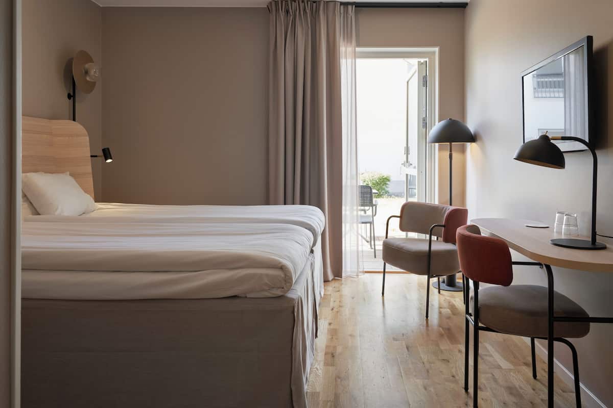 Ett hotellrum med en säng, två stolar, ett skrivbord, en lampa, en spegel och ett fönster med gardiner.