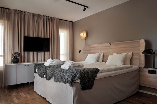 A modern hotel room with a large bed, a flat-screen TV mounted on the wall, a bedside table with a lamp, and a window with curtains. - Quality Hotel Visby (Visby)