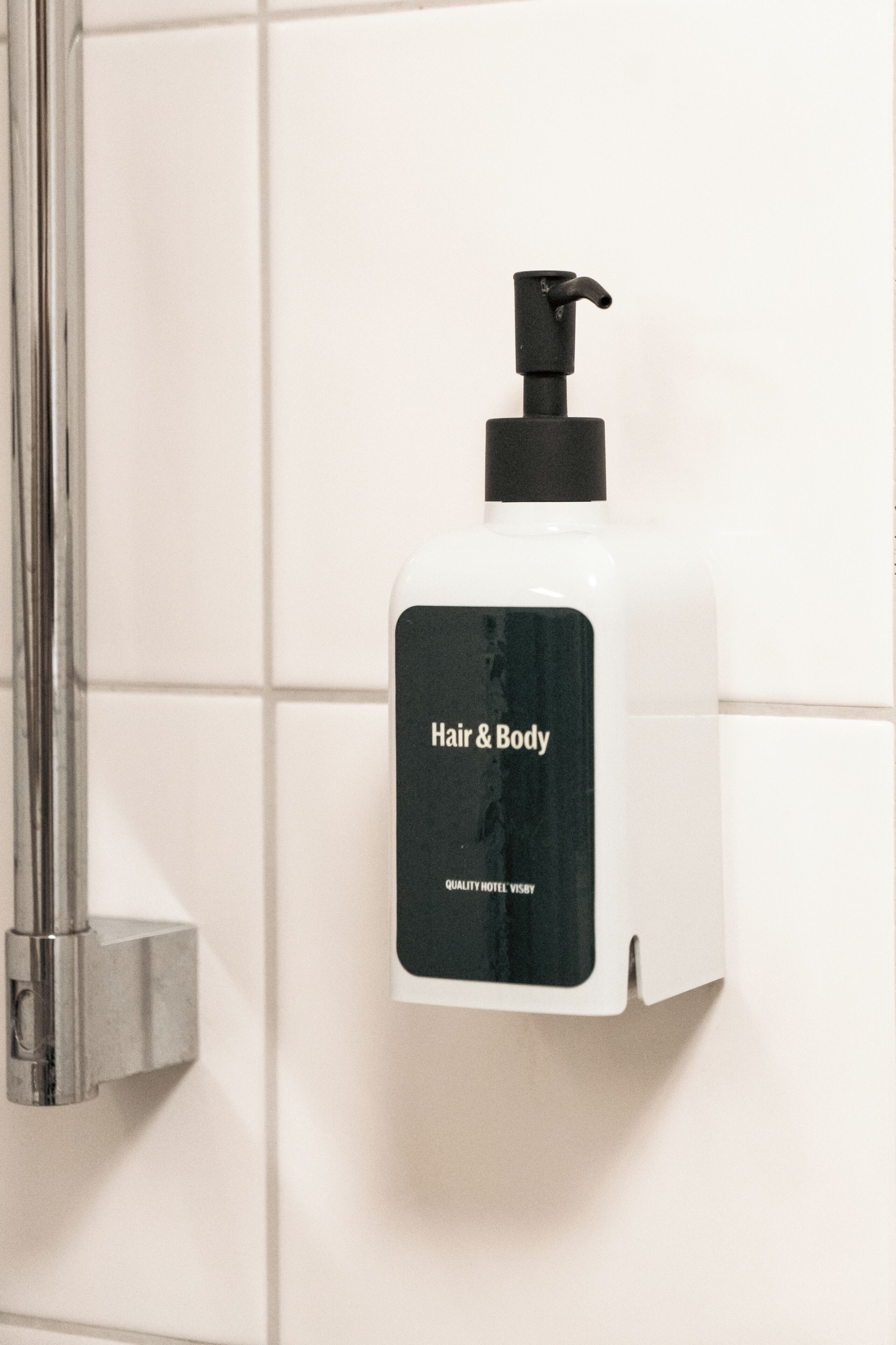 A white bottle of Hair & Body soap with a black pump, mounted on a white tiled wall.