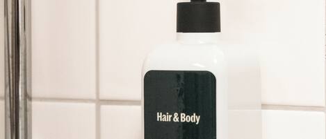 A white bottle of Hair & Body soap with a black pump, mounted on a white tiled wall.