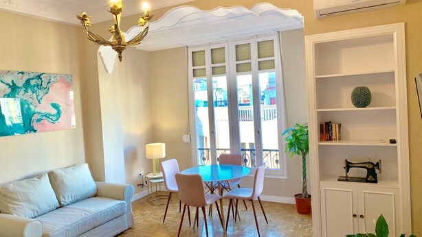 A living room with a sofa, a dining table with chairs, a chandelier, a large window with a view, a bookshelf, and a potted plant. - Hi Valencia Cánovas (Valencia)