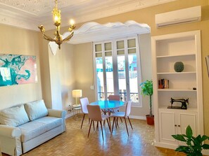 Una sala de estar con un sofá, una mesa de comedor con sillas, una araña, un ventanal con vistas, una estantería y una planta en maceta.
