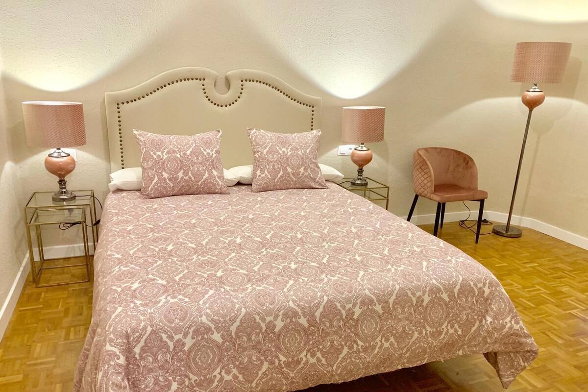 A neatly made bed with a patterned bedspread, two matching pillows, and two bedside lamps on matching nightstands. A chair is positioned near the bed.