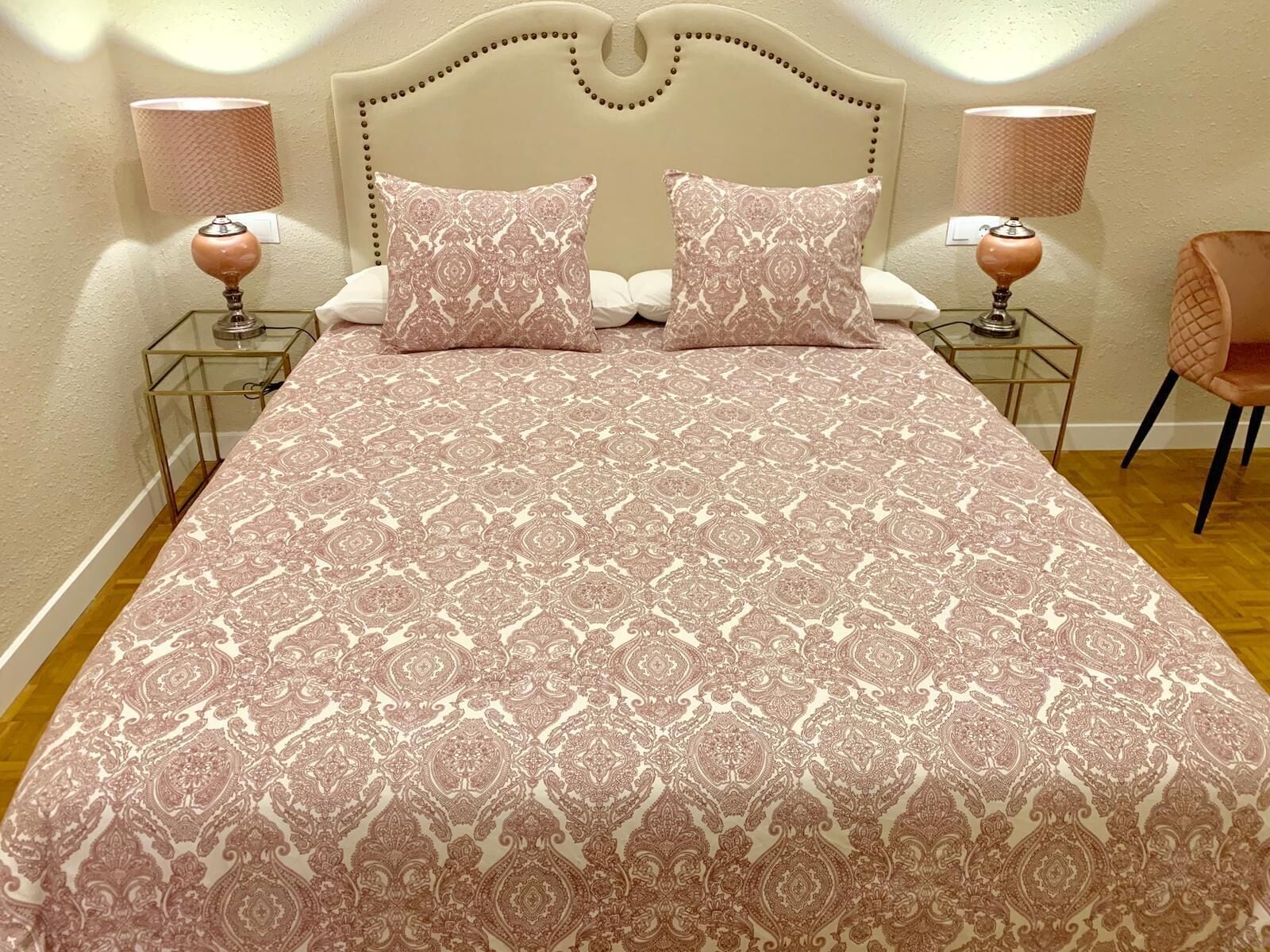 A bed with a patterned bedspread, two matching pillows, two bedside lamps, and a chair.
