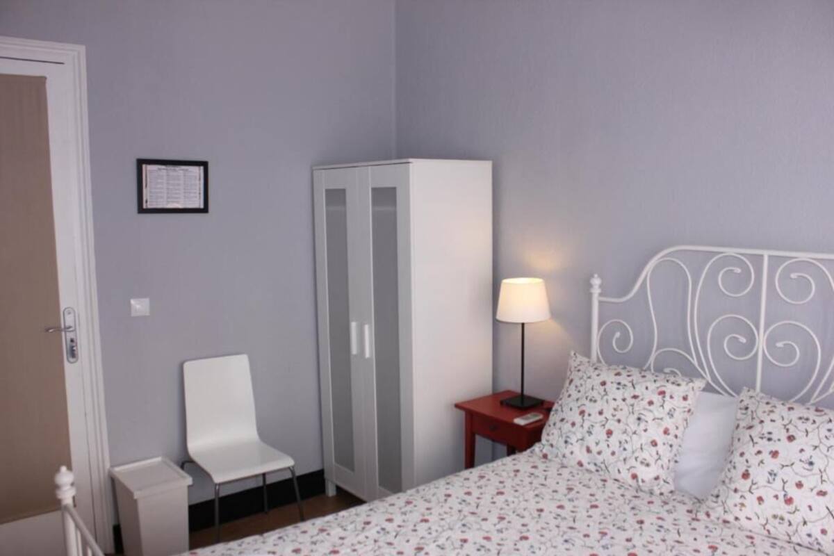 A bedroom with a bed, a chair, a wardrobe, and a nightstand with a lamp.