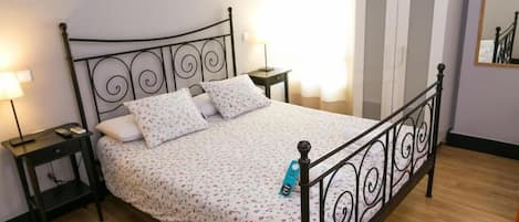 A bed with a metal frame, a bedside table with a lamp, and a wardrobe in a room with wooden flooring.