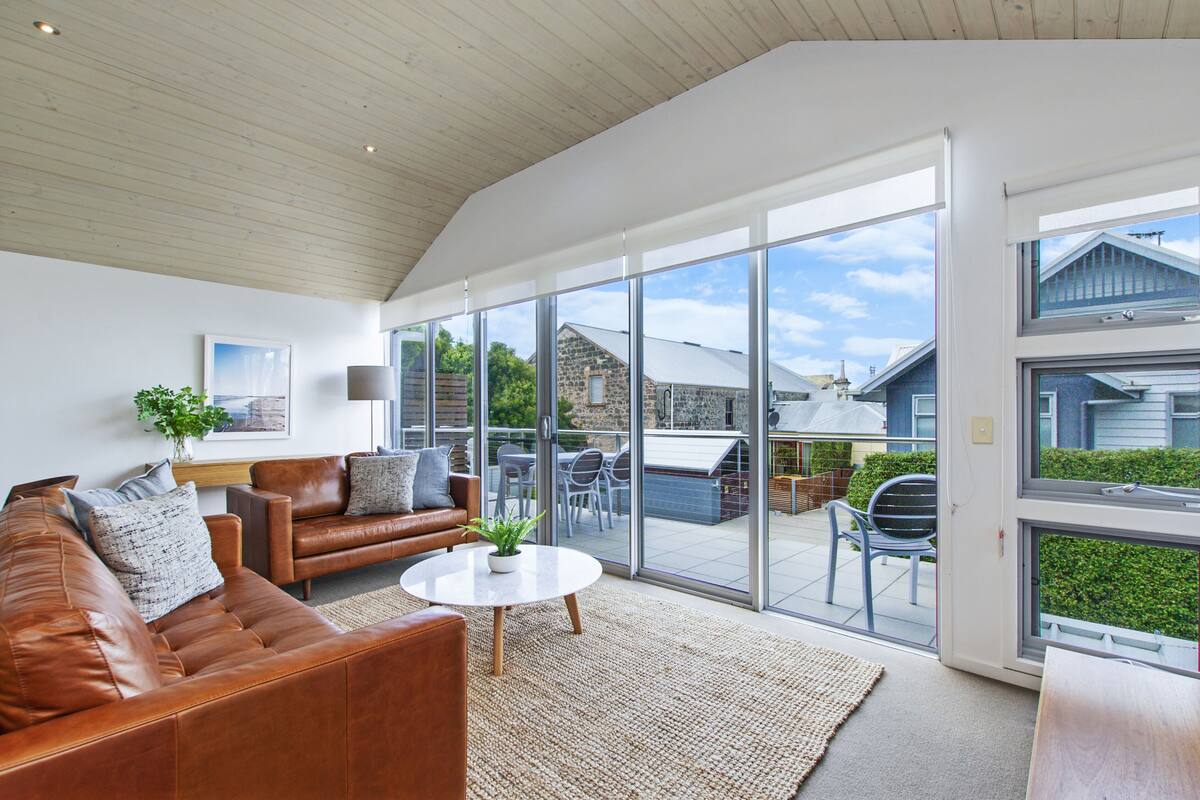 A spacious living room with a leather sofa, a coffee table, and a large sliding glass door leading to an outdoor patio with a table and chairs.
