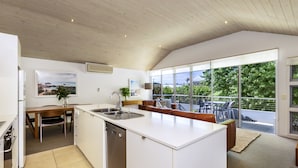 Une cuisine moderne dotée d’un grand îlot central, un coin repas avec une table et des chaises en bois, et un balcon offrant une vue sur la verdure.