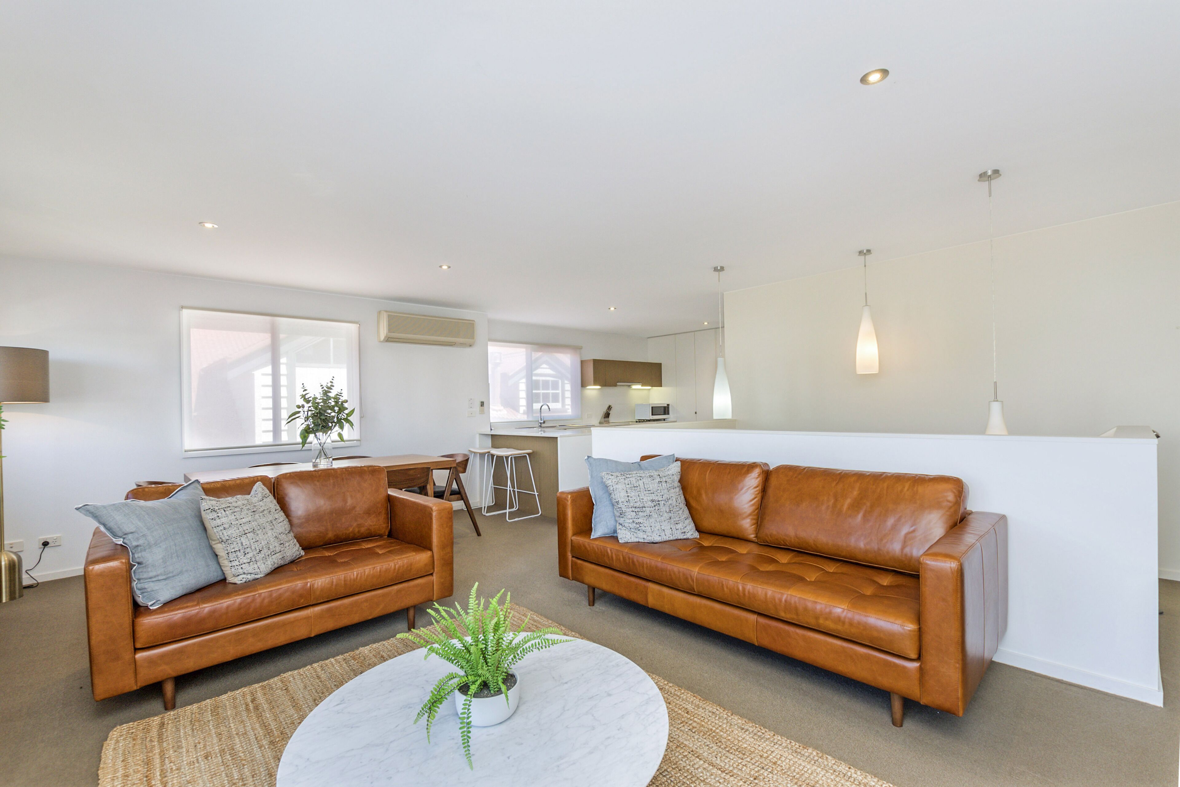A modern living room with two brown leather sofas, a white coffee table with a plant, and a kitchen area in the background.