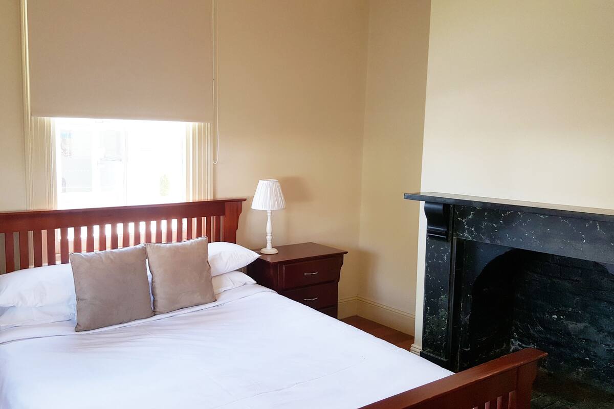 A neatly made bed with white linens and two pillows, a bedside table with a lamp, a wooden nightstand, and a fireplace.