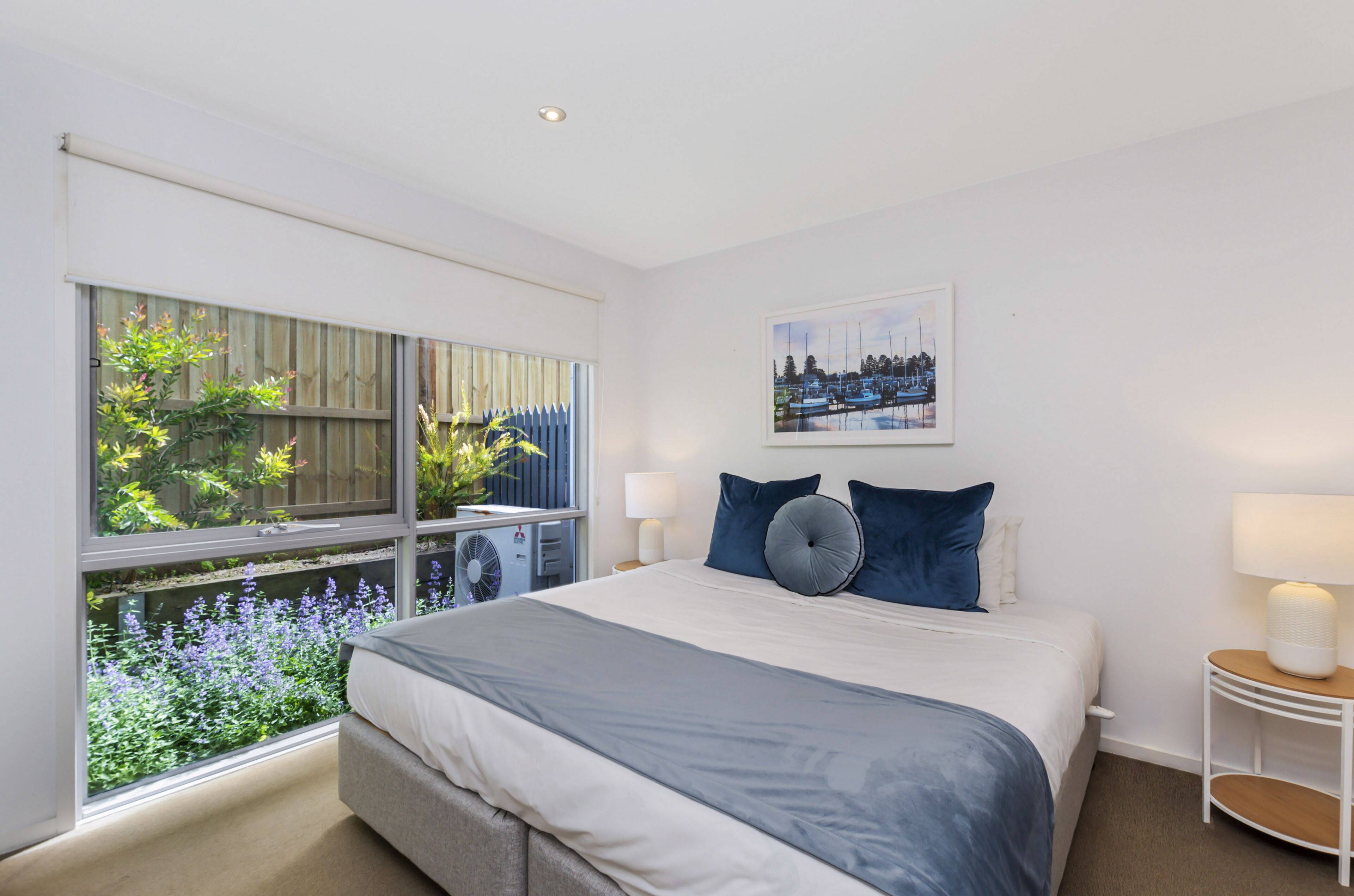 A bedroom with a large window, a bed, two bedside tables with lamps, and a framed picture on the wall.