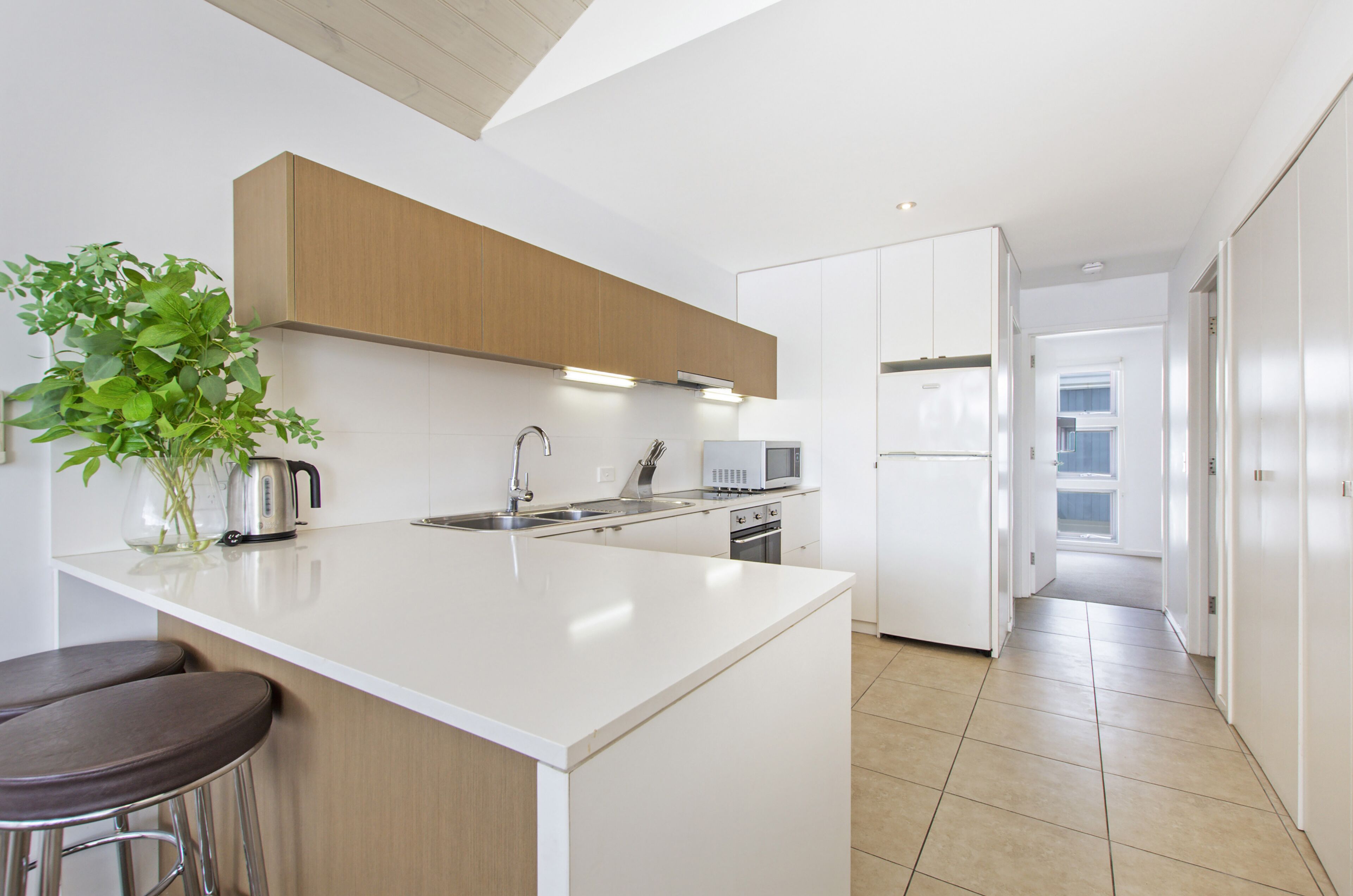 A modern kitchen with white cabinetry, a central island with bar stools, a stainless steel kettle, and a potted plant.