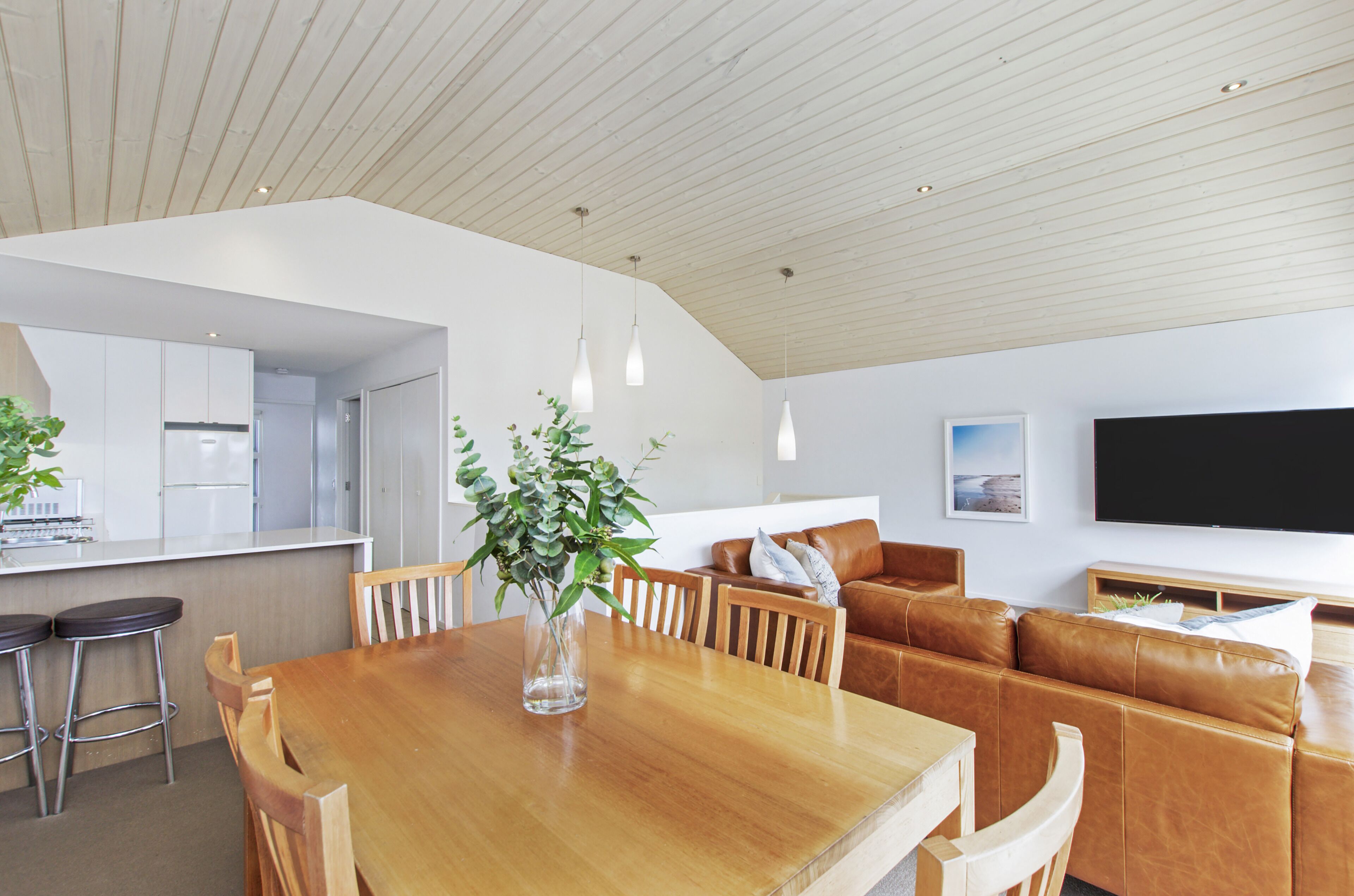 A modern kitchen with white cabinets, a wooden dining table, and bar stools. There is a large window with a view of the outdoors.