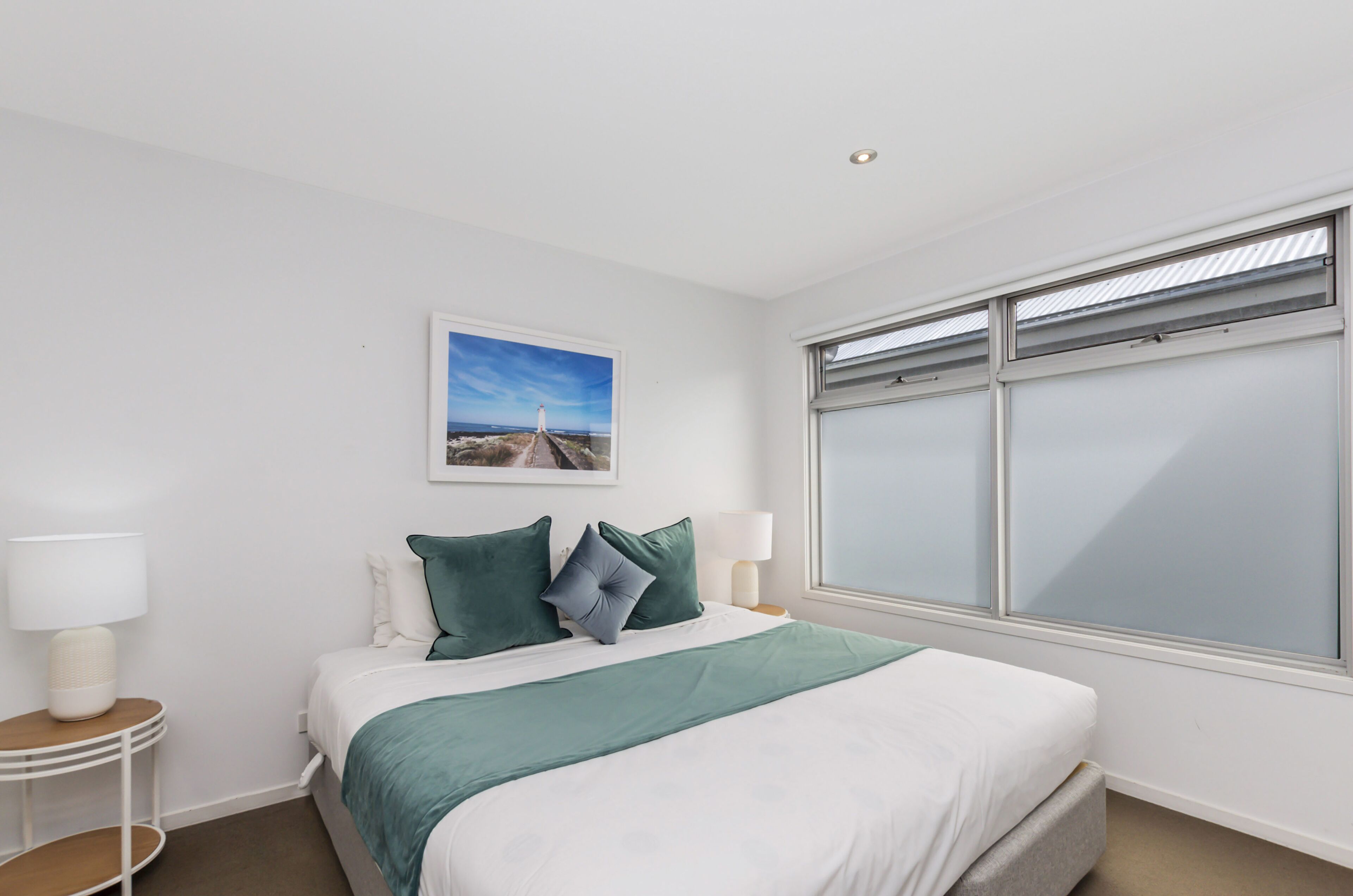 Une chambre à coucher bien rangée, avec un lit, deux tables de chevet dotées de lampes, une fenêtre à vitre dépolie et un tableau encadré accroché au mur.