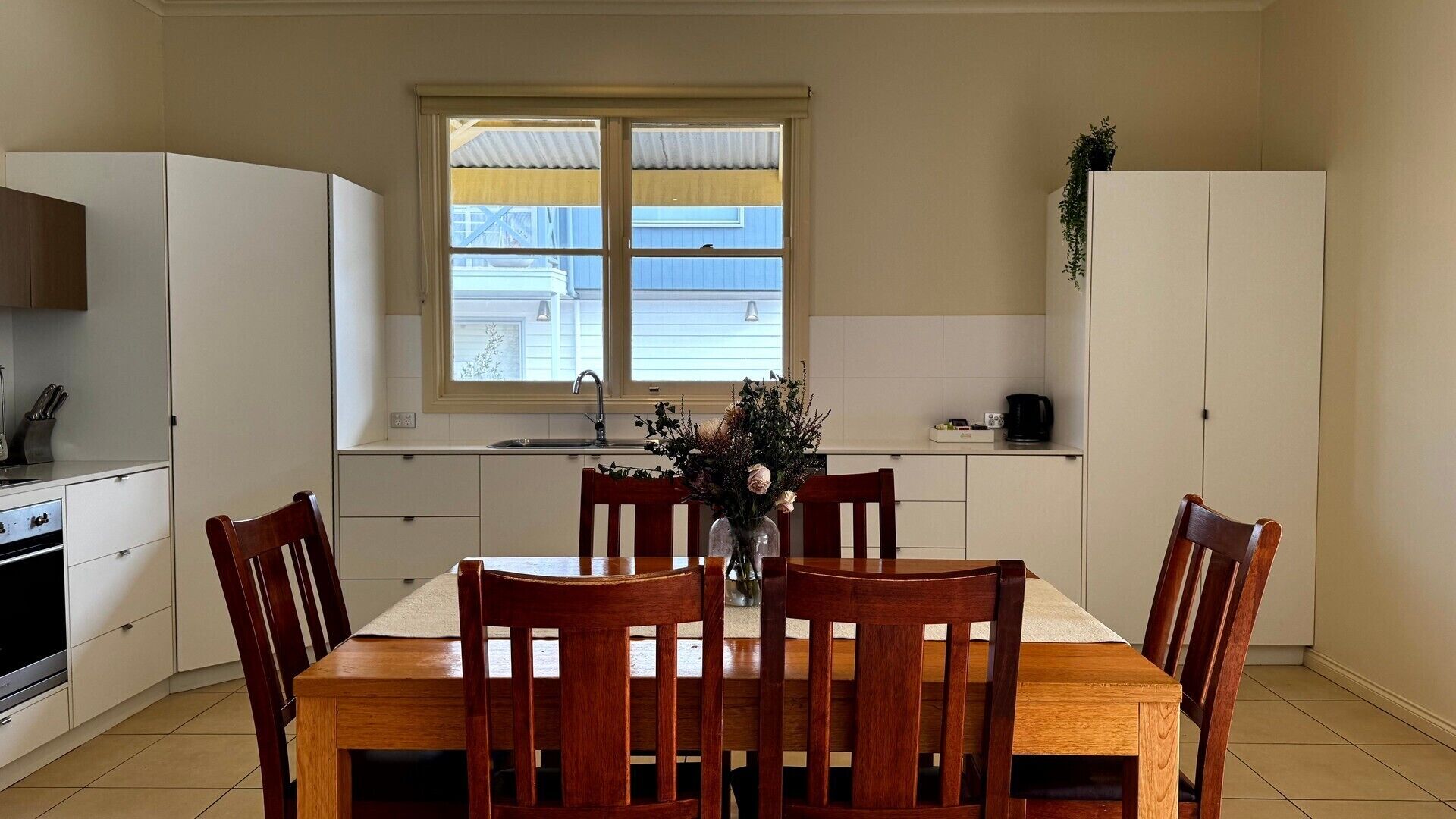 A dining area with a wooden table and chairs, a vase with flowers, and a window with blinds.