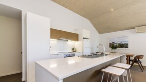 Une cuisine moderne avec un plan de travail blanc, des armoires en bois et une cuisinière encastrée. On y trouve deux tabourets de bar et une petite table à manger avec des chaises.