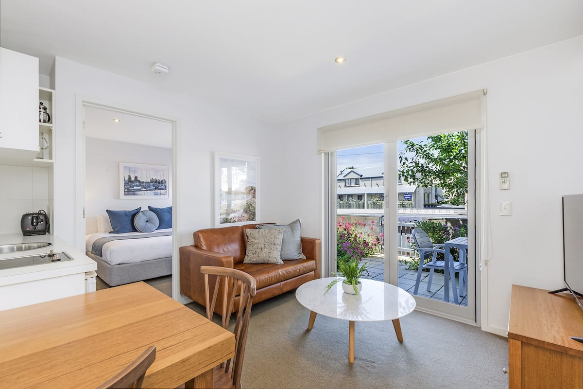A modern apartment with a kitchenette, a dining area, a living room with a sofa and a coffee table, and a sliding glass door leading to a balcony with outdoor seating.